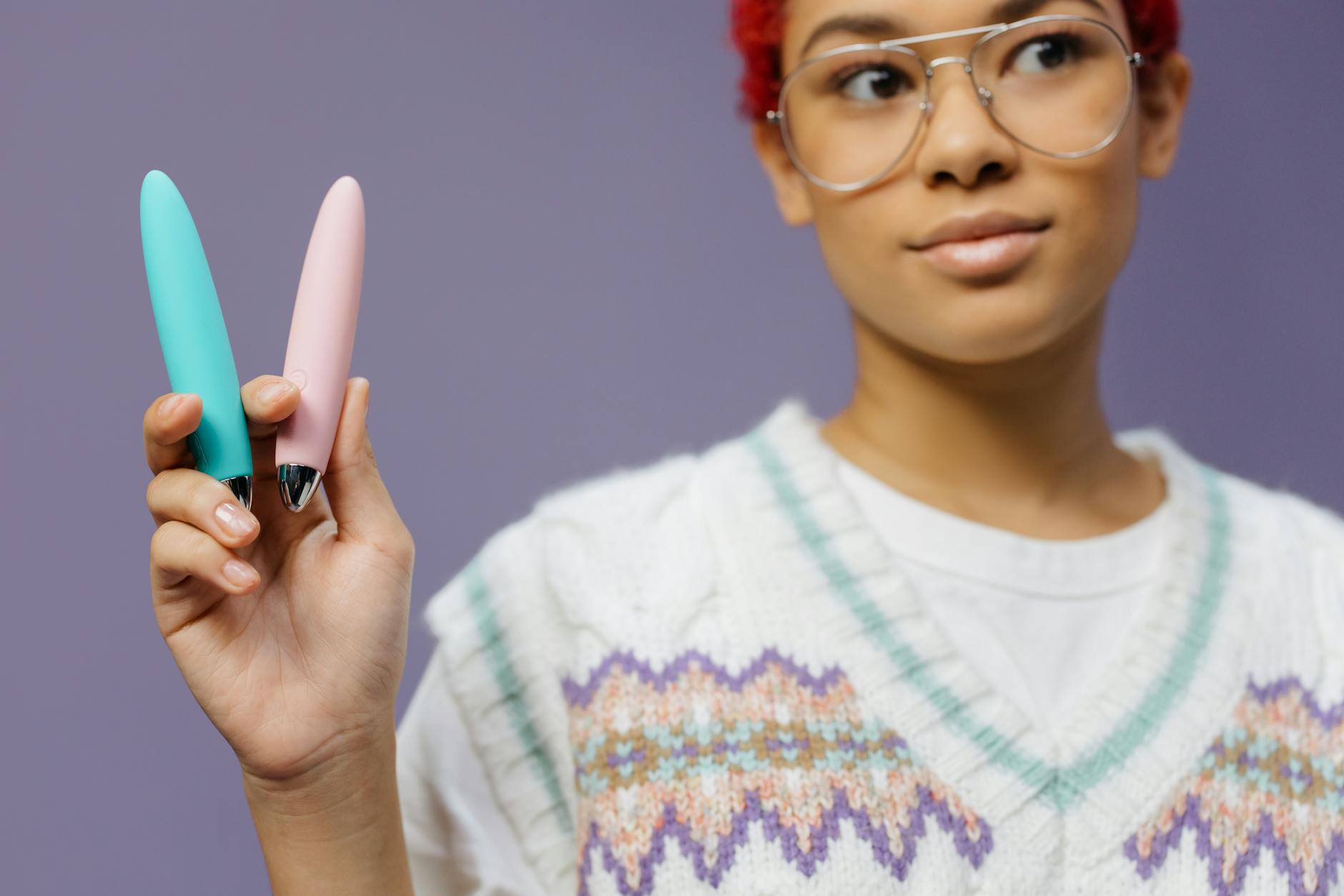 紫色の背景で眼鏡をかけた若い女性が2つのカラフルなバイブレーターを持っている様子